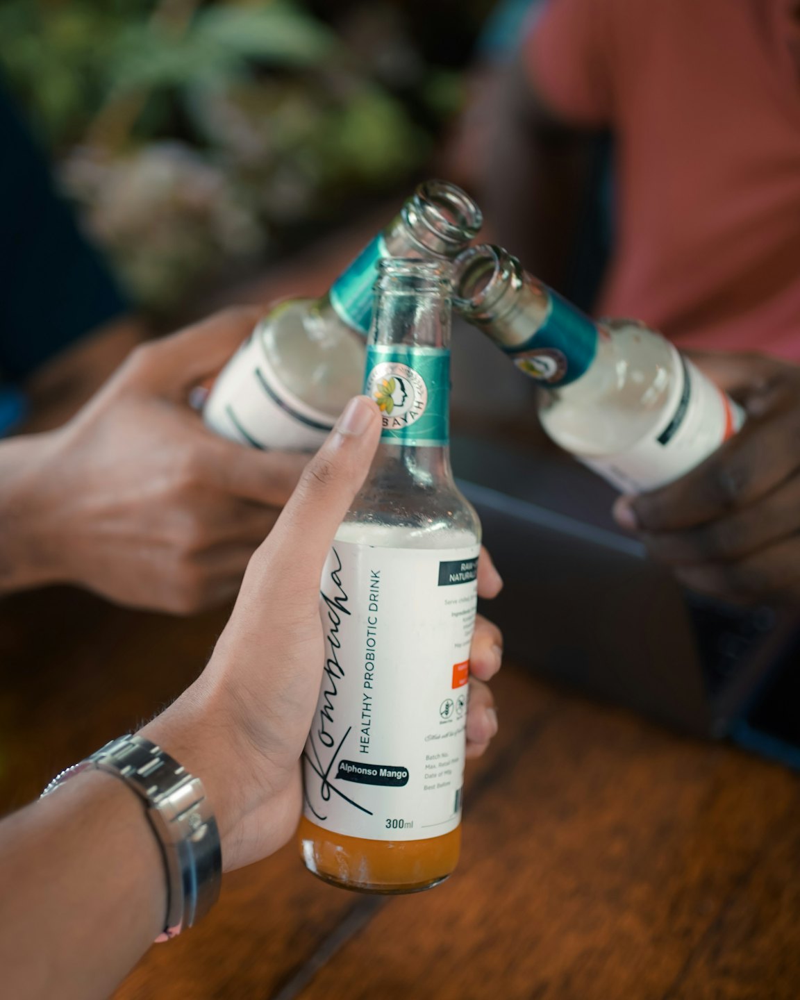 Tres personas brindan con botellas de mango kombucha sobre una mesa.